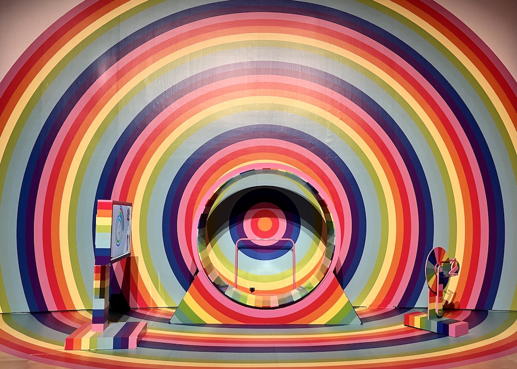 A rainbow coloured life-size hamster wheel against a background of circular rainbows