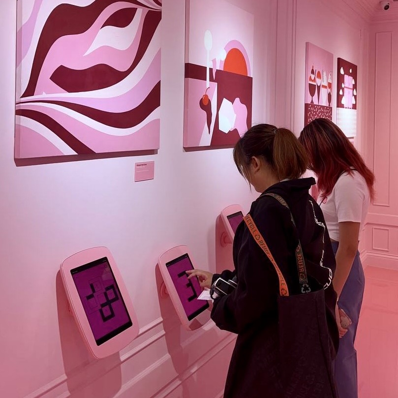 Two people playing games on a tablet mounted to a pink wall filled with paintings